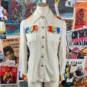 May include: A cream-colored linen shirt with rainbow trim on the cuffs and shoulders. The shirt has a button-down collar and two flap pockets.