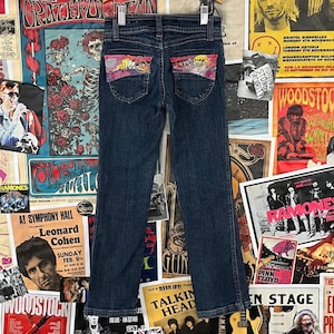 May include: Dark wash denim jeans with decorative patches on the back pockets. The patches feature colorful designs with a sunset theme. The jeans are hanging against a backdrop of vintage concert posters.