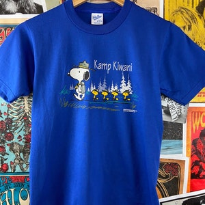 K&ouml;nnte beinhalten: Ein k&ouml;nigsblaues T-Shirt mit einem Aufdruck von Snoopy, der einen Rangerhut tr&auml;gt und einen Rucksack tr&auml;gt. Der Text "Kamp Kiwani" ist in Wei&szlig; &uuml;ber Snoopy gedruckt.