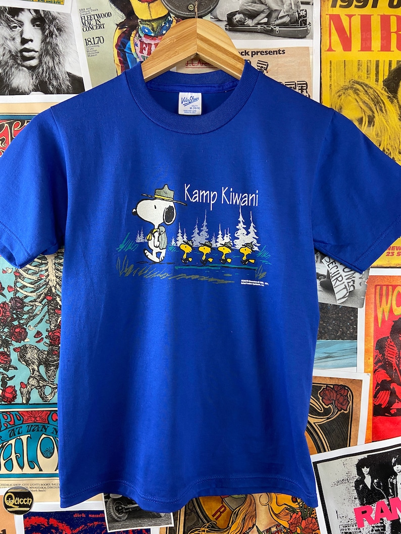 K&ouml;nnte beinhalten: Ein k&ouml;nigsblaues T-Shirt mit einem Aufdruck von Snoopy, der einen Rangerhut tr&auml;gt und einen Rucksack tr&auml;gt. Der Text "Kamp Kiwani" ist in Wei&szlig; &uuml;ber Snoopy gedruckt.