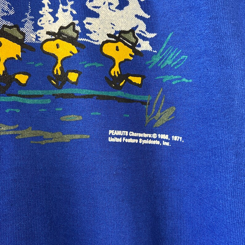 K&ouml;nnte beinhalten: Blauer Pullover mit einem Aufdruck von drei Zeichentrickfiguren der Peanuts, die Rangerh&uuml;te tragen und in einer Reihe laufen. Der Text "PEANUTS Characters: &copy; 1958, 1971, United Feature Syndicate, Inc." ist auf der Unterseite des Pullovers aufgedruckt.