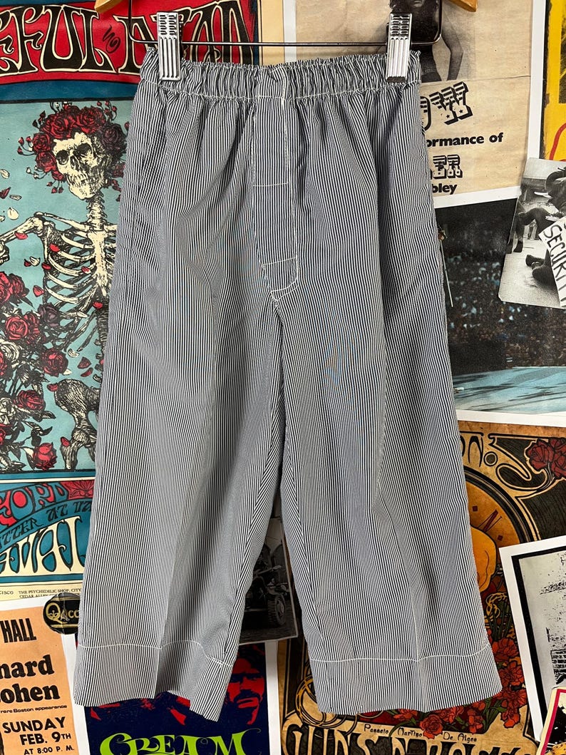May include: Blue and white striped cropped pants with an elastic waistband. The pants have a vertical stripe pattern and are made of a lightweight fabric. The hem of the pants is finished with a white border. The pants are hanging on a hanger.