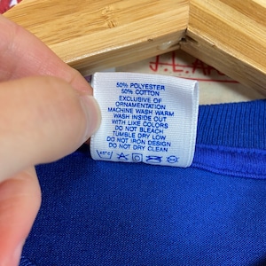 K&ouml;nnte beinhalten: Ein blaues Pflegeetikett mit wei&szlig;em Text, der lautet: "50% Polyester 50% Baumwolle Ausgenommen Verzierungen Maschinenw&auml;sche warm Auf links waschen Mit &auml;hnlichen Farben waschen Nicht bleichen Trocknergeeignet bei niedriger Temperatur Nicht auf das Design b&uuml;geln Nicht chemisch reinigen 40'c"