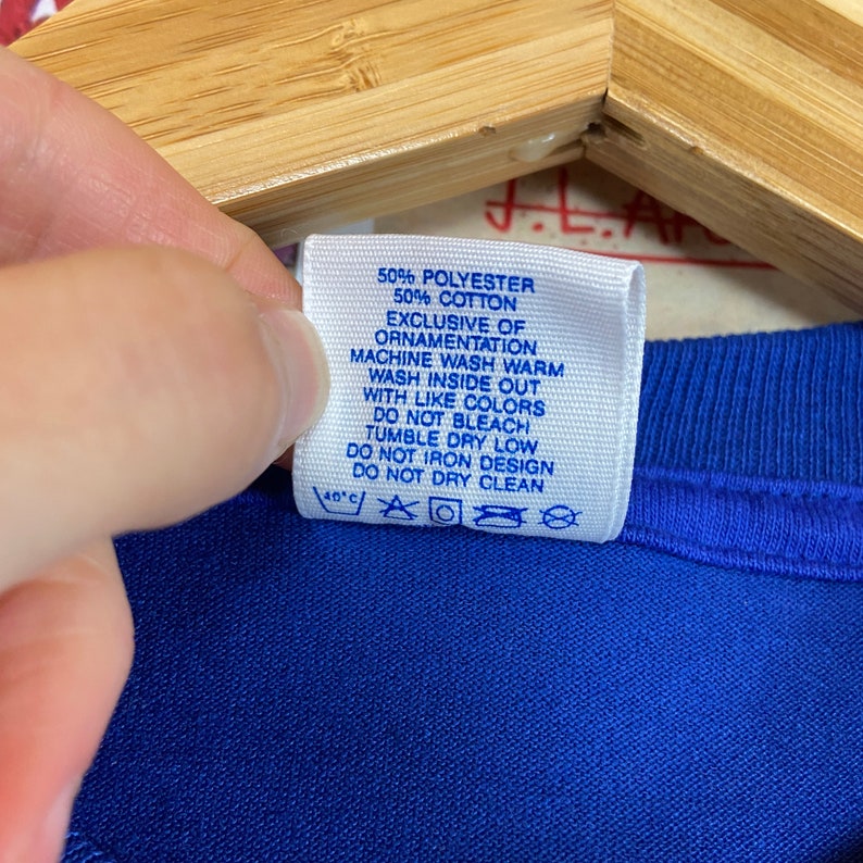 K&ouml;nnte beinhalten: Ein blaues Pflegeetikett mit wei&szlig;em Text, der lautet: "50% Polyester 50% Baumwolle Ausgenommen Verzierungen Maschinenw&auml;sche warm Auf links waschen Mit &auml;hnlichen Farben waschen Nicht bleichen Trocknergeeignet bei niedriger Temperatur Nicht auf das Design b&uuml;geln Nicht chemisch reinigen 40'c"