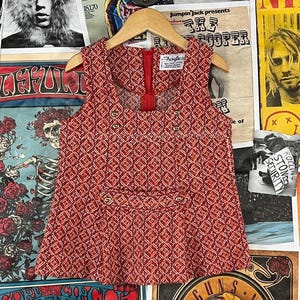 May include: A sleeveless red top with a geometric diamond pattern, a red zip, and gold buttons. The top is hanging on a wooden hanger. The background features vintage posters.