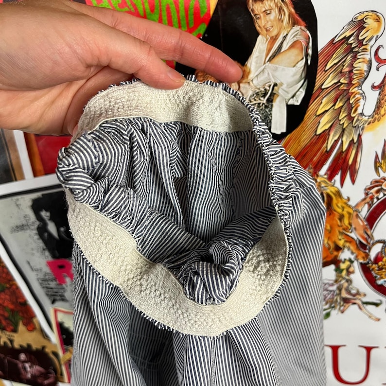 May include: Close-up of a blue and white striped garment. The interior lining features a similar striped pattern. The cuff is trimmed with a cream-colored, textured fabric. The garment is held by a hand.