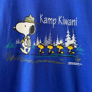 K&ouml;nnte beinhalten: Blaues T-Shirt mit einem Cartoon-Bild von Snoopy, der einen Scout-Hut tr&auml;gt und einen Rucksack tr&auml;gt. Der Text "Kamp Kiwani" ist auf dem T-Shirt aufgedruckt. F&uuml;nf kleinere Cartoon-Bilder von Snoopy befinden sich unter dem Hauptbild.