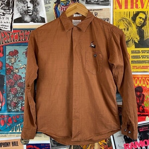 May include: A brown long-sleeved button-down shirt with a small embroidered design on the left chest pocket.