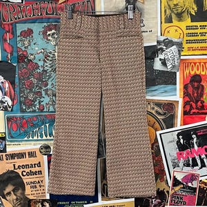 May include: Tan and brown patterned pants with a wide leg. The pants are hanging against a backdrop of vintage concert posters and album art, including The Ramones and Woodstock.