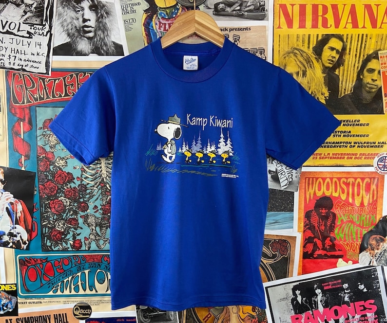K&ouml;nnte beinhalten: Ein k&ouml;nigsblaues T-Shirt mit einem Aufdruck von Snoopy, der einen Rangerhut tr&auml;gt und einen Rucksack tr&auml;gt. Der Text "Kamp Kiwani" ist &uuml;ber Snoopy gedruckt.