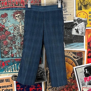 May include: Blue pinstripe pants hanging on a hanger. The pants have a high waist and a slight flare at the bottom. The background features vintage concert posters and artwork.