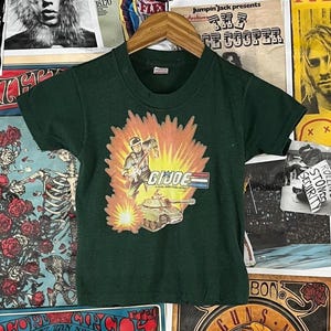 May include: Dark green vintage t-shirt featuring a graphic of a soldier, tank, and the text "G.I. Joe" in a red, white, and blue design. The shirt is short-sleeved and has a crew neck.