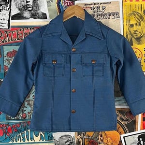 Puede incluir: Una chaqueta vaquera azul vintage con cuello, dos bolsillos en el pecho y botones. La chaqueta tiene botones marrones y detalles de costura visibles. La chaqueta se muestra en una percha de madera.
