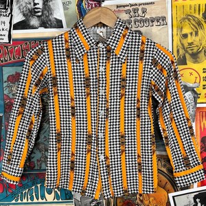 Może przedstawiać: Koszula z długim rękawem zapinana na guziki, w czarno-biały wzór w kratę i pomarańczowe paski. Koszula ma kołnierzyk i zapięcie na guziki.