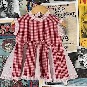May include: A red and white gingham dress with a bow at the waist and eyelet lace trim. The dress is hanging on a wooden hanger against a backdrop of vintage posters.