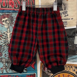 May include: Red, black, and green plaid pants with a gathered, balloon-style leg and black cuffs. The pants are hanging on a metal hanger. The fabric has a classic tartan pattern.
