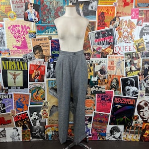 May include: Gray tweed pants with a button fly and pleats. The pants are on a mannequin against a background of vintage concert posters.