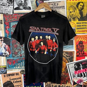 May include: Black t-shirt featuring a graphic from Star Trek V: The Final Frontier. The design shows the cast in red uniforms within a white circle, with the film's title above. The shirt is displayed on a wooden hanger against a backdrop of vintage posters.