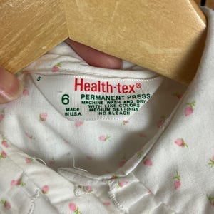 K&ouml;nnte beinhalten: Wei&szlig;es Baumwollhemd mit kleinem rosa Blumendruck. Das Hemd hat einen Kragen und einen Knopfverschluss. Das Etikett lautet "Health-tex&reg; Permanent Press Machine Wash and Dry with Like Colors Medium Settings No Bleach Made in U.S.A."