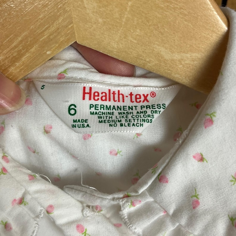 K&ouml;nnte beinhalten: Wei&szlig;es Baumwollhemd mit kleinem rosa Blumendruck. Das Hemd hat einen Kragen und einen Knopfverschluss. Das Etikett lautet "Health-tex&reg; Permanent Press Machine Wash and Dry with Like Colors Medium Settings No Bleach Made in U.S.A."