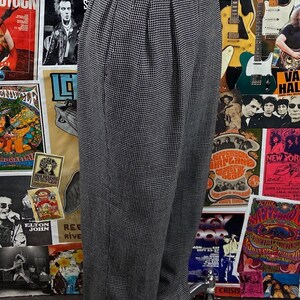 Pode incluir: Cal&ccedil;a xadrez preta e branca de cintura alta e com pregas. A cal&ccedil;a est&aacute; em um manequim e &eacute; exibida contra um fundo de cartazes de concertos vintage.