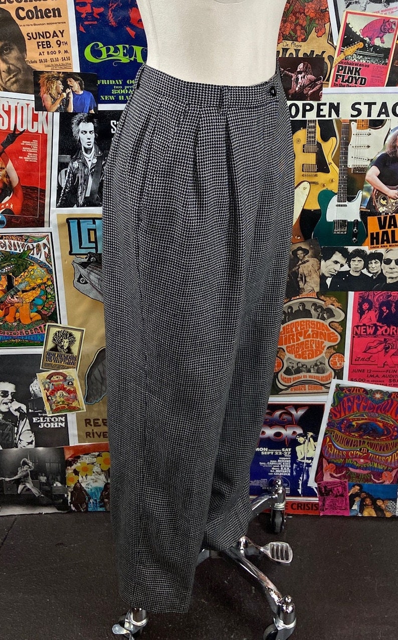 Pode incluir: Cal&ccedil;a xadrez preta e branca de cintura alta e com pregas. A cal&ccedil;a est&aacute; em um manequim e &eacute; exibida contra um fundo de cartazes de concertos vintage.