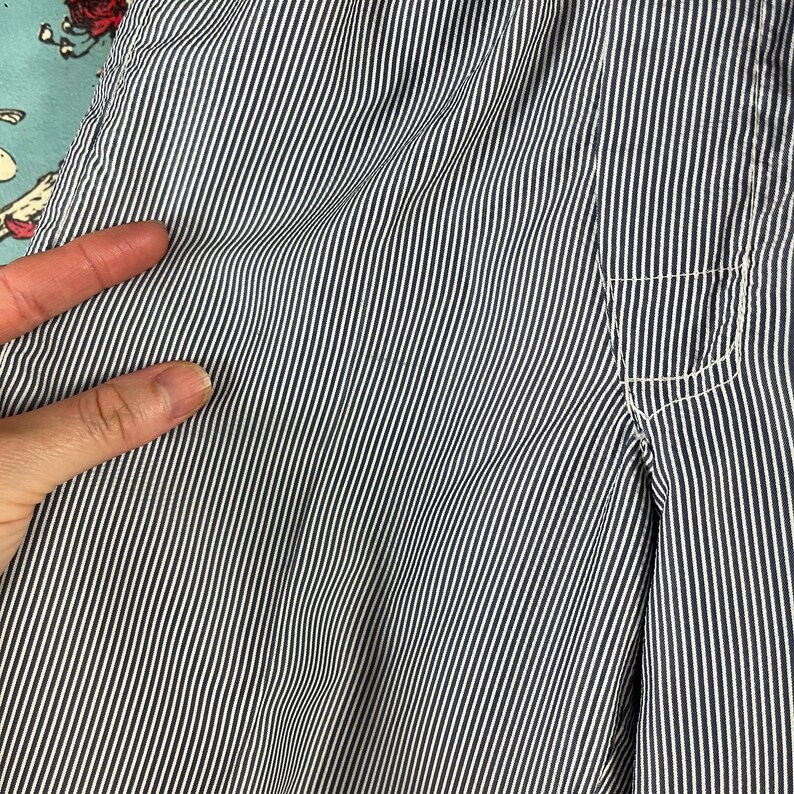 May include: Close-up of blue and white striped fabric. The fabric has a vertical stripe pattern with a pocket detail on the right side. The fabric is slightly wrinkled, suggesting it is made of a soft material.