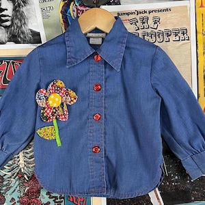 May include: A blue denim shirt with a red flower applique. The flower has a yellow center and is made of different fabrics. The shirt has a button-up front and long sleeves.
