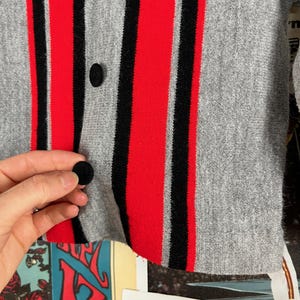 Puede incluir: Primer plano de un c&aacute;rdigan a rayas grises, rojas y negras con botones negros. El c&aacute;rdigan est&aacute; hecho de una tela suave y de punto.