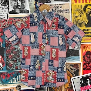 Könnte beinhalten: Ein blaues und rotes kurzärmeliges Hemd mit Knöpfen und einem Patchwork-Muster mit einem sich wiederholenden Muster von kleinen Mädchen in Kleidern. Das Hemd hat einen weißen Kragen und eine Knopfleiste.