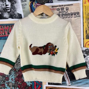 May include: A cream-colored knitted sweater with a brown and white embroidered dog and flowers. The sweater has brown, green, and white stripes around the cuffs and hem.