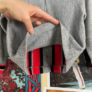 Puede incluir: Sudadera gris con pu&ntilde;os a rayas rojas y negras. La sudadera tiene una cremallera.