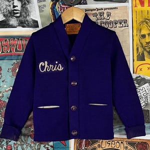 Peut inclure: Un cardigan bleu foncé avec un col châle et une fermeture boutonnée. Le nom "Chris" est brodé en écriture blanche sur la poitrine. Il comporte deux poches avant avec une bordure blanche. Le cardigan est présenté sur un cintre en bois.