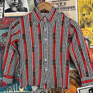 May include: A long-sleeved button-up shirt with a black and white houndstooth pattern and red vertical stripes. The shirt has a pointed collar and a button-up front.