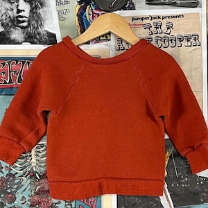 Puede incluir: Una sudadera de color óxido, de manga larga, con cuello redondo y puños acanalados. La parte superior para niños se exhibe en una percha de madera sobre un fondo de carteles vintage. La sudadera está hecha de una tela suave y texturizada.
