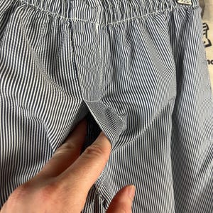 May include: Close-up of blue and white striped fabric, possibly clothing. The fabric has a vertical stripe pattern. A hand is reaching into a pocket on the garment. The fabric is gathered at the top.