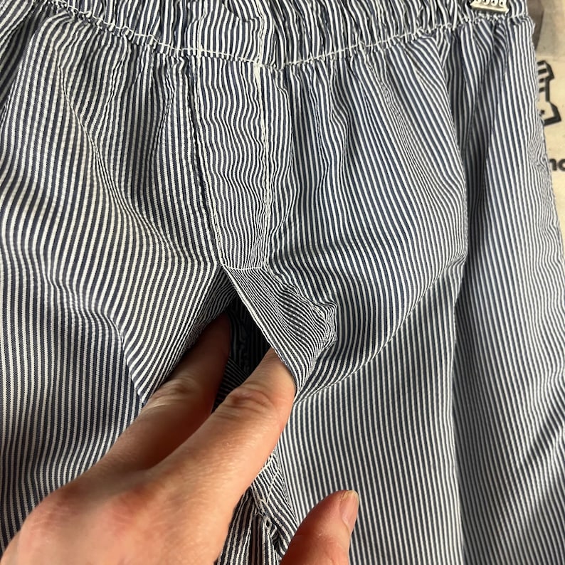 May include: Close-up of blue and white striped fabric, possibly clothing. The fabric has a vertical stripe pattern. A hand is reaching into a pocket on the garment. The fabric is gathered at the top.