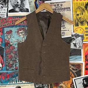 May include: Brown corduroy vest with a V-neck, button closure, and two front pockets. The vest is displayed on a wooden hanger against a backdrop of vintage concert posters.
