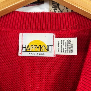 Może przedstawiać: Czerwony sweter dzianinowy z ż&oacute;łtym logo słońca i tekstem "HAPPYKNIT" oraz "MADE IN U.S.A." na białej metce. Metka zawiera r&oacute;wnież instrukcje pielęgnacyjne: 100% akryl, pranie w pralce w ciepłej wodzie, suszenie w suszarce bębnowej w niskiej temperaturze, nie wybielać, wyprodukowano w USA, WPL 6816.