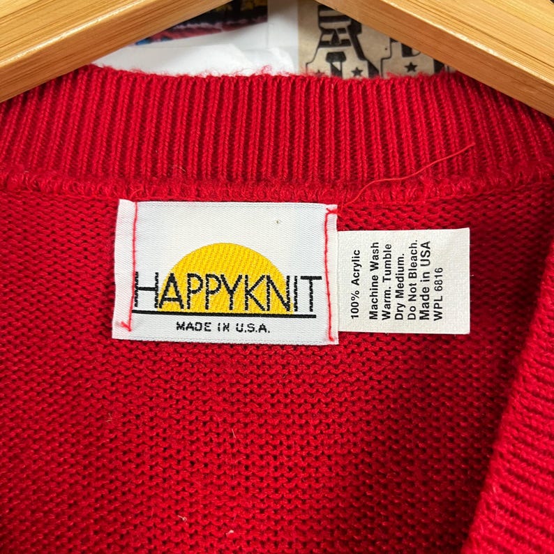 Może przedstawiać: Czerwony sweter dzianinowy z ż&oacute;łtym logo słońca i tekstem "HAPPYKNIT" oraz "MADE IN U.S.A." na białej metce. Metka zawiera r&oacute;wnież instrukcje pielęgnacyjne: 100% akryl, pranie w pralce w ciepłej wodzie, suszenie w suszarce bębnowej w niskiej temperaturze, nie wybielać, wyprodukowano w USA, WPL 6816.