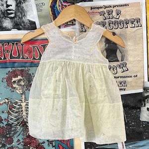 May include: A vintage, pale yellow baby dress with a delicate, sheer overlay. The dress features a simple, sleeveless design with a round neckline, small pockets, and embroidered floral accents. It hangs on a wooden hanger against a backdrop of vintage posters.