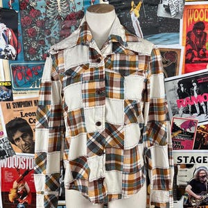 May include: A patchwork button-down shirt with a white collar and cuffs. The shirt features a mix of plaid and solid patches in shades of brown, cream, and blue. The shirt is displayed against a backdrop of vintage concert posters.