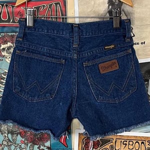 May include: Dark blue denim shorts with a frayed hem. The shorts feature two back pockets with a signature Wrangler stitching design and a brown leather Wrangler patch. The shorts are displayed on a hanger.