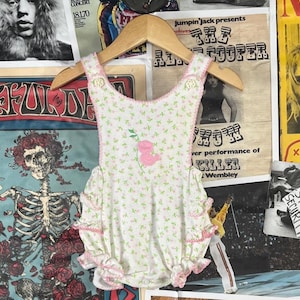 May include: White baby romper with a floral pattern and pink trim, featuring a pink rose applique. The romper is hanging on a wooden hanger against a backdrop of vintage concert posters.