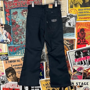 May include: Dark navy blue flared jeans with a classic five-pocket design. The jeans have a high waist and a small label on the back pocket. The background features vintage concert posters.