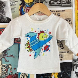 Puede incluir: Camiseta blanca de manga larga con un gráfico colorido de un dinosaurio de dibujos animados en un cohete con el texto "U.S.A."