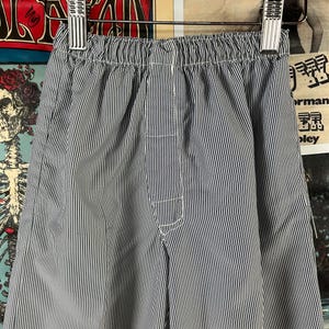 May include: Close-up of blue and white striped pants hanging on a metal hanger. The pants have an elastic waistband and white stitching details. The fabric has a vertical stripe pattern.