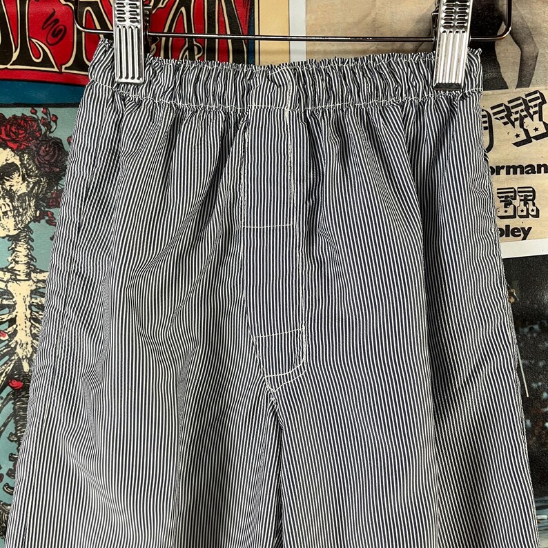 May include: Close-up of blue and white striped pants hanging on a metal hanger. The pants have an elastic waistband and white stitching details. The fabric has a vertical stripe pattern.