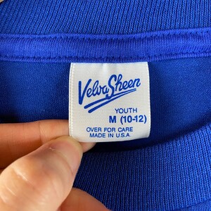 K&ouml;nnte beinhalten: Ein wei&szlig;es Etikett mit blauem Text, auf dem in kursiver Schrift "Velva Sheen" steht. Unter dem Markennamen steht "Youth M (10-12)" und "Over for care Made in U.S.A."