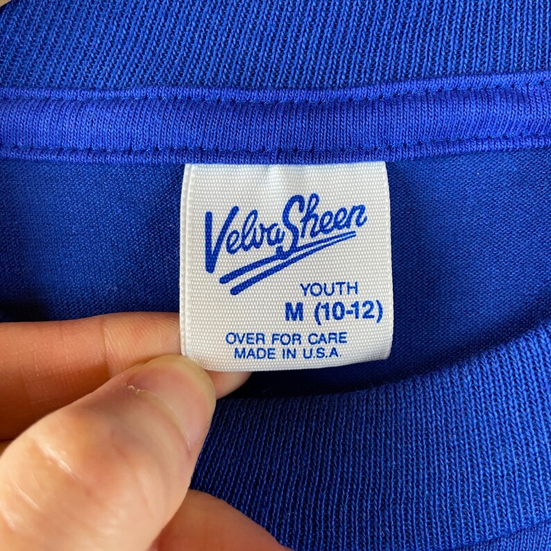 K&ouml;nnte beinhalten: Ein wei&szlig;es Etikett mit blauem Text, auf dem in kursiver Schrift "Velva Sheen" steht. Unter dem Markennamen steht "Youth M (10-12)" und "Over for care Made in U.S.A."